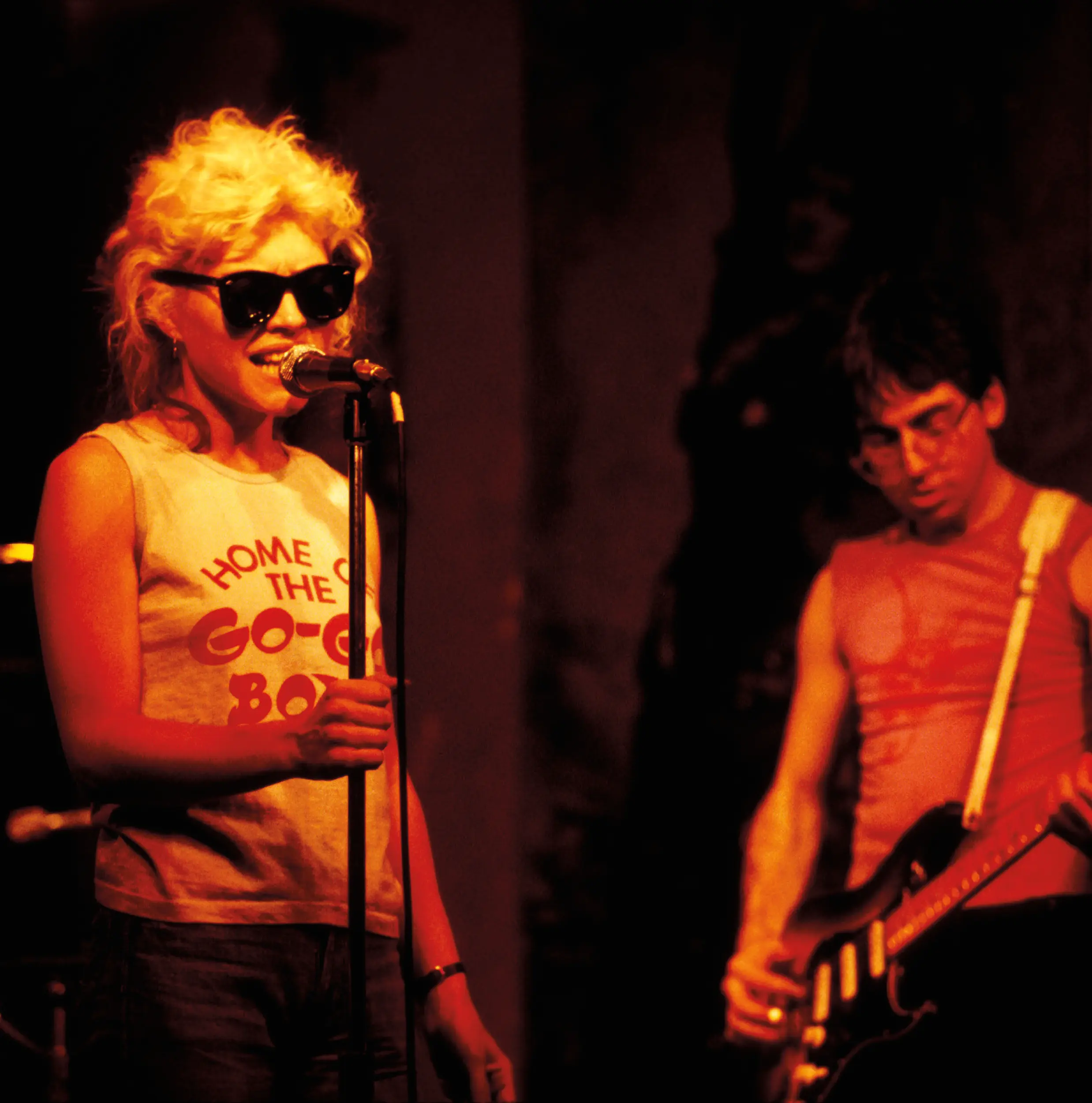 A blonde woman wearing sunglasses and a sleeveless shirt sings into a microphone on stage, while a man in a red shirt plays an electric guitar beside her, both bathed in warm, orange stage lighting.